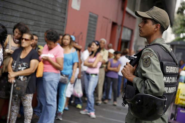 A Venezuelan soldier stands guard next to people forming a line to try to buy cornmeal flour and margarine at a pharmacy in Caracas
